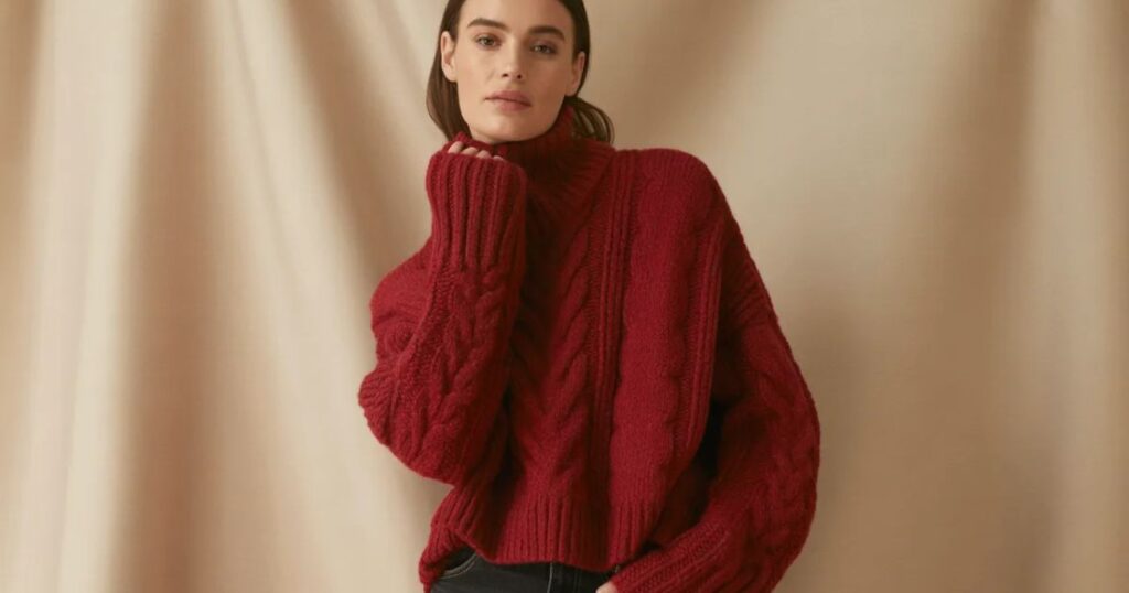 Model seated on a chair wearing a loose Red Sweater Fashion turtleneck sweater and black jeans in a minimalist studio.